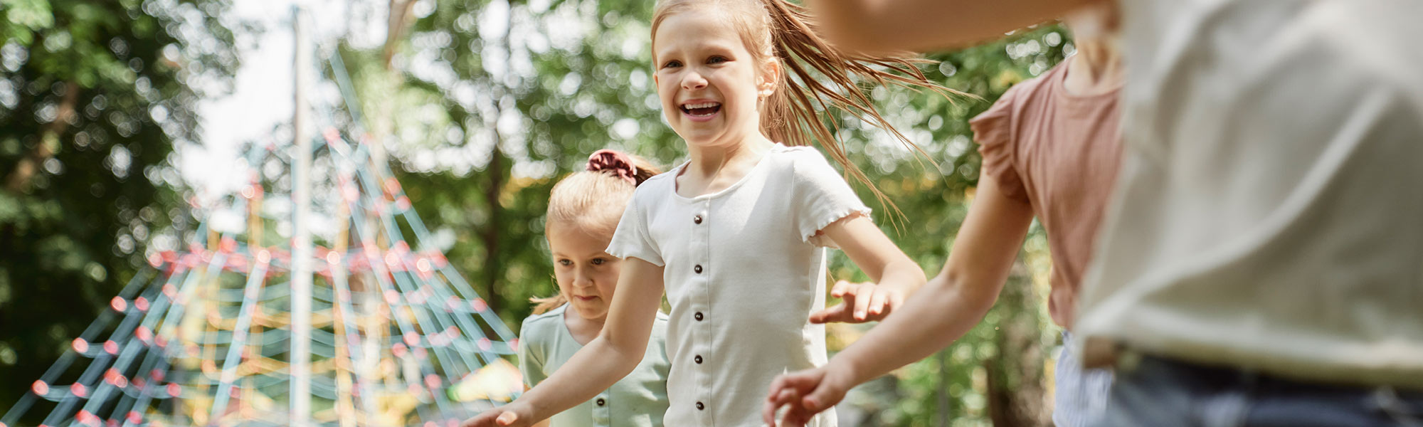 Mehrere Kinder spielen glücklich im Freien bei sonnigem Wetter, im Hintergrund ist ein Klettergerüst zu sehen.